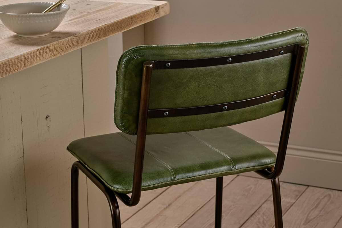 A nkuku Ukari Counter Chair in rich green leather, featuring a padded seat and backrest, sits by a light wood counter. On the wooden floor nearby, a white bowl with spoon complements the room’s neutral walls.