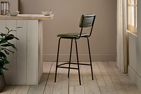The nkuku Ukari Counter Chair - Rich Green, featuring a tall green leather seat and metal frame, stands by a light wood kitchen island in a bright room with wood floors, beige walls, a corner plant, and sunlight through sheer curtains.