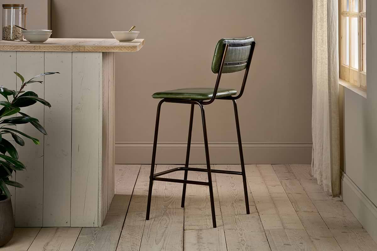 The nkuku Ukari Counter Chair - Rich Green, featuring a tall green leather seat and metal frame, stands by a light wood kitchen island in a bright room with wood floors, beige walls, a corner plant, and sunlight through sheer curtains.