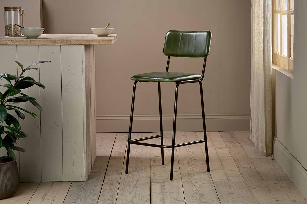 The Ukari Counter Chair - Rich Green by nkuku, featuring a metal frame, stands on light wooden flooring next to a beige kitchen island with a potted plant and bowls, as soft sunlight streams through sheer curtains to the right.