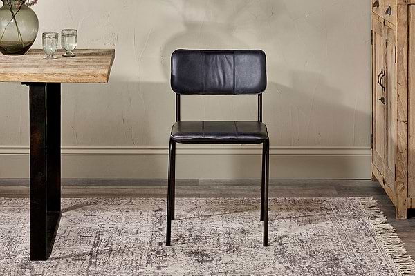 The Ukari Leather Dining Chair - Aged Black by nkuku, featuring buffalo leather upholstery and a metal frame, is placed on a gray patterned rug between a wooden table with glassware and a wooden cabinet against a light beige wall.
