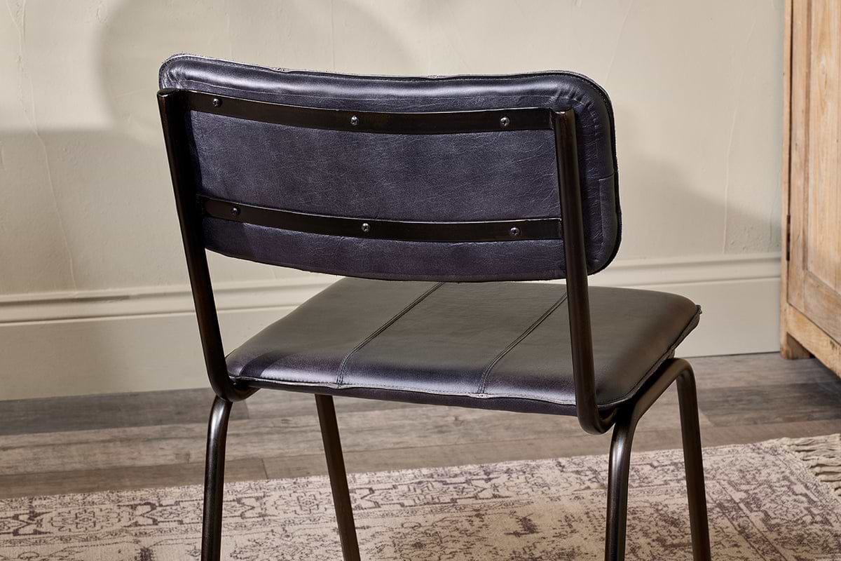The Ukari Leather Dining Chair - Aged Black by nkuku is shown from behind, featuring a black metal frame and padded buffalo leather upholstery. It sits on a patterned rug over a wooden floor, with beige walls and wood furniture in the background.