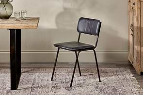 The Ukari Leather Dining Chair - Aged Black by nkuku, with a metal frame and buffalo leather upholstery, sits on a patterned rug near a wooden table set with glasses and a vase. The indoor setting features textured beige walls and a wooden cabinet.