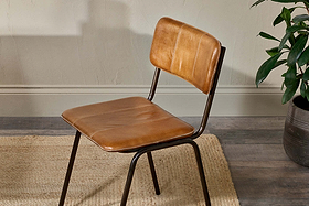 The nkuku Ukari Leather Dining Chair in aged tan, featuring padded buffalo leather upholstery and slim black metal legs, sits on a light woven rug atop a wood floor near a textured light wall, with a green potted plant partly visible to the right.