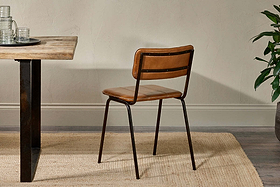 The nkuku Ukari Leather Dining Chair in aged tan features brown buffalo leather upholstery and a black metal frame, placed on a beige woven rug next to a wooden table with clear glasses, a tray, and a green potted plant by a cream wall.