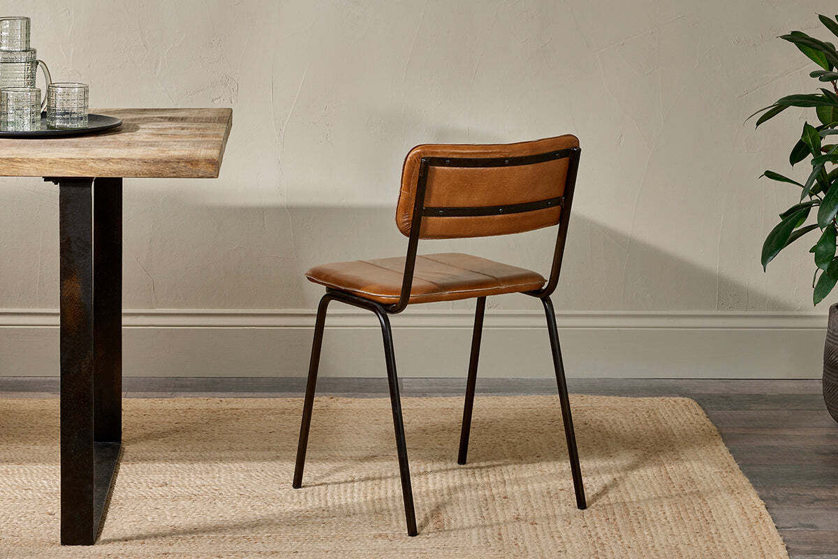 The nkuku Ukari Leather Dining Chair in aged tan features brown buffalo leather upholstery and a black metal frame, placed on a beige woven rug next to a wooden table with clear glasses, a tray, and a green potted plant by a cream wall.