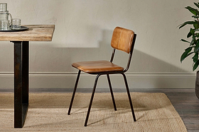 The nkuku Ukari Leather Dining Chair in Aged Tan, featuring cushioned leather upholstery and black metal legs, stands by a wooden table on a beige rug. A plant appears on the right against a neutral wall.
