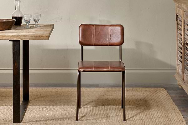 The Ukari Leather Dining Chair in chocolate brown by nkuku, with a metal frame, stands on a woven beige rug. To the left is a rustic table set with glassware and a bowl, against a light textured wall and wooden furniture on the right.