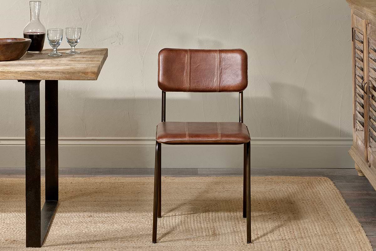 The Ukari Leather Dining Chair in chocolate brown by nkuku, with a metal frame, stands on a woven beige rug. To the left is a rustic table set with glassware and a bowl, against a light textured wall and wooden furniture on the right.