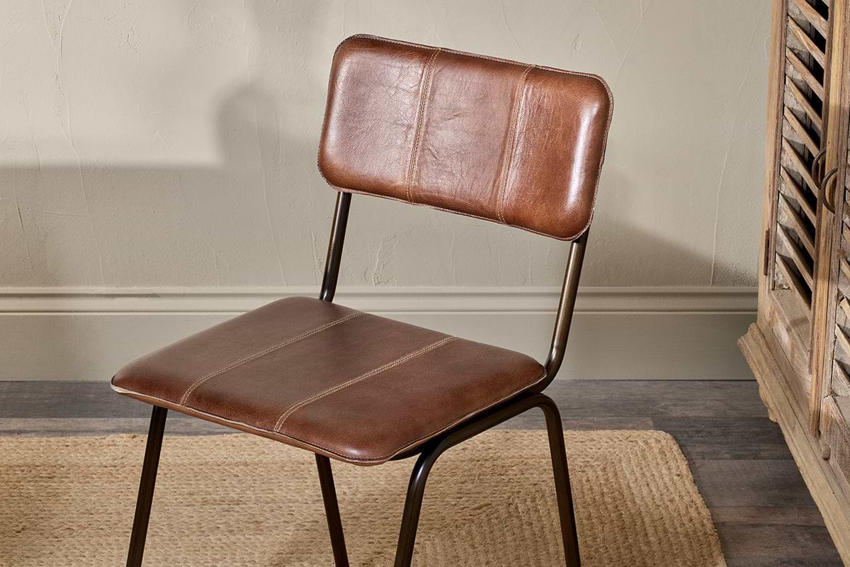 The nkuku Ukari Leather Dining Chair in Chocolate Brown with stitched detailing and a metal frame sits on a woven rug. Beside it, a distressed wooden cabinet complements the rich buffalo leather upholstery.