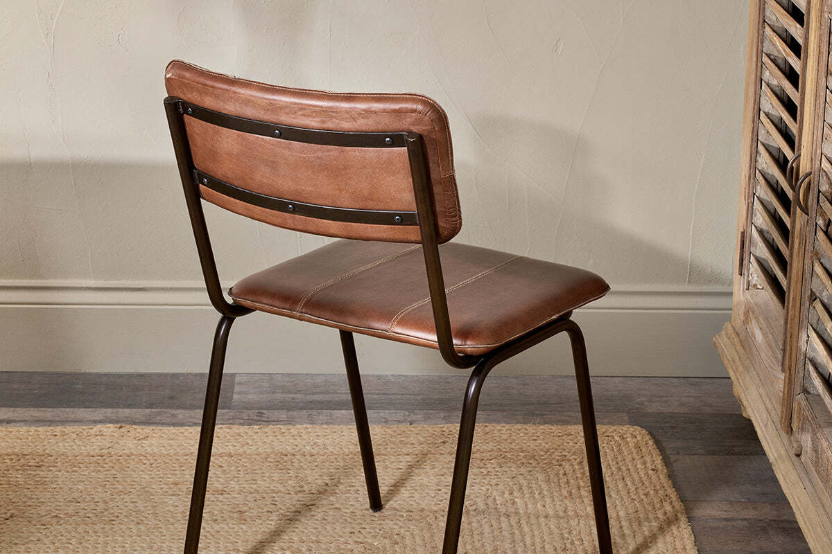 The nkuku Ukari Leather Dining Chair in chocolate brown features buffalo leather upholstery, a cushioned seat and backrest, and black metal legs. It’s styled indoors on a beige woven rug beside wooden furniture and a textured taupe wall.