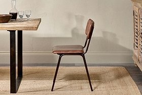 The nkuku Ukari Leather Dining Chair in chocolate brown, featuring a metal frame and padded leather seat, sits on a woven rug beside a wooden dining table topped with glasses, a decanter, and bowl, set by a textured beige wall and cabinet.