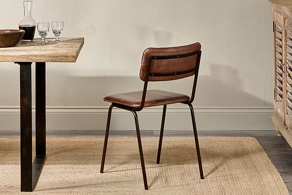 The Ukari Leather Dining Chair in chocolate brown by nkuku, with buffalo leather upholstery and a metal frame, sits on a woven rug beside a wooden dining table set with glasses, bowl, and plate in a softly lit room with neutral walls.