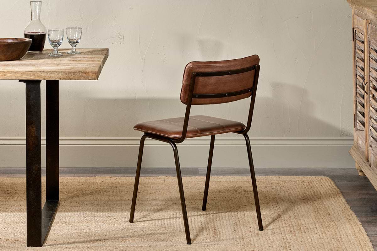 The Ukari Leather Dining Chair in chocolate brown by nkuku, with buffalo leather upholstery and a metal frame, sits on a woven rug beside a wooden dining table set with glasses, bowl, and plate in a softly lit room with neutral walls.