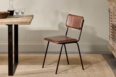 The nkuku Ukari Leather Dining Chair - Chocolate Brown, with padded seat and backrest, stands on a woven beige rug by a wooden table set for dining, bringing comfort and style to the warmly lit rustic interior.