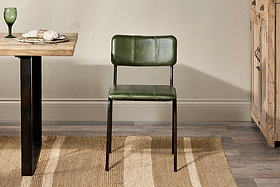 A nkuku Ukari Leather Dining Chair in Rich Green, featuring buffalo leather and a metal frame, sits on a striped rug by a wooden table set with a green glass, napkin, and plate; to its right is a rustic wooden cabinet.