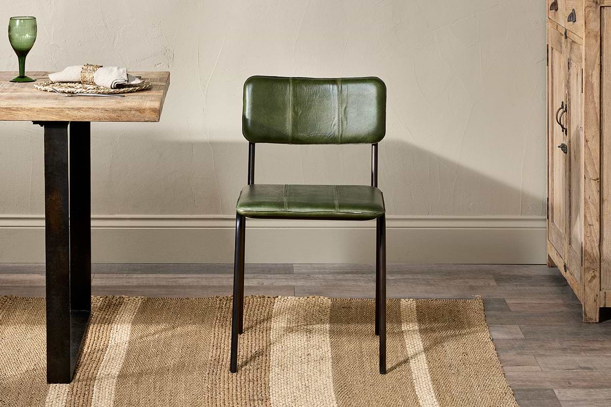 A nkuku Ukari Leather Dining Chair in Rich Green, featuring buffalo leather and a metal frame, sits on a striped rug by a wooden table set with a green glass, napkin, and plate; to its right is a rustic wooden cabinet.