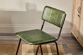 The Ukari Leather Dining Chair by nkuku, in rich green buffalo leather with a metal frame and visible stitching, sits on a woven rug. The setting features wood flooring, a cream wall, and a wooden cabinet.