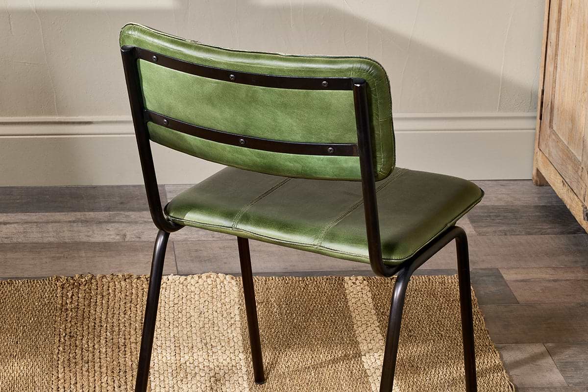 The Ukari Leather Dining Chair in Rich Green by nkuku, featuring a black metal frame, sits on a tan woven rug atop wooden floors. The stitched leather upholstery is visible from behind, with beige walls and wooden furniture in the background.
