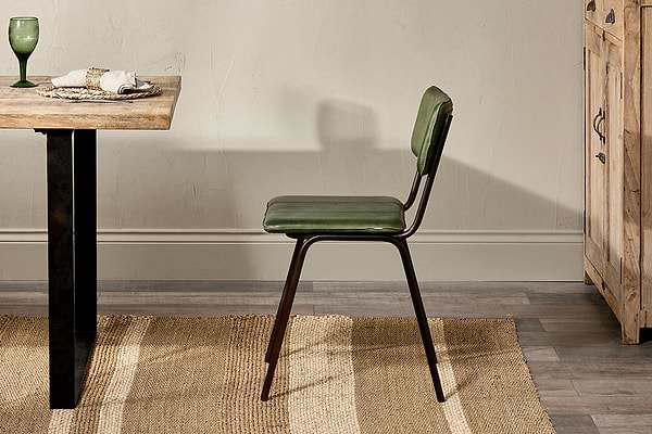 A nkuku Ukari Leather Dining Chair in rich green with metal legs stands by a wooden dining table set with a green glass, plates, and napkins. The indoor scene includes a woven rug, wooden floor, beige wall, and a wooden cabinet on the right.