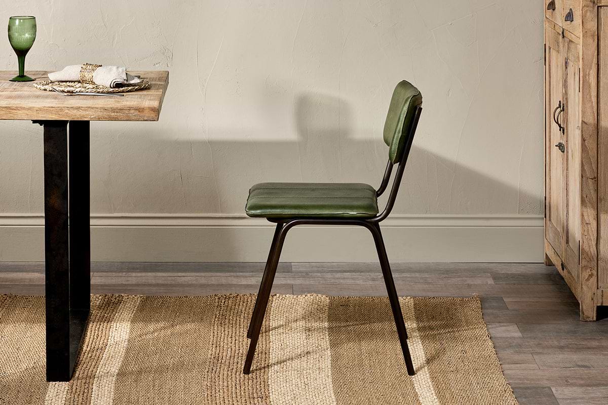 A nkuku Ukari Leather Dining Chair in rich green with metal legs stands by a wooden dining table set with a green glass, plates, and napkins. The indoor scene includes a woven rug, wooden floor, beige wall, and a wooden cabinet on the right.