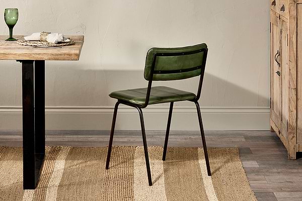 The Ukari Leather Dining Chair in rich green by nkuku, with its dark metal frame, sits beside a wooden table on a striped rug. A green glass on the table complements the chair's elegant style.