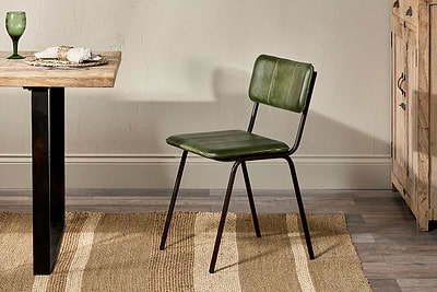 The Ukari Leather Dining Chair by nkuku in rich green buffalo leather with a metal frame sits on a striped rug near a wooden dining table set with a green glass, dishes, and napkin. A light wall and wooden cabinet are visible in the background.