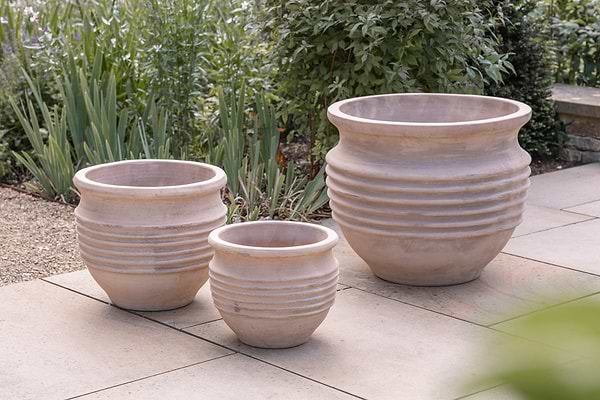 Three Ullu Terracotta Planters by nkuku, in natural terracotta with a ribbed design, sit on a stone patio amid lush greenery. These unglazed planters suit both indoor and outdoor use, adding rustic charm to any space.