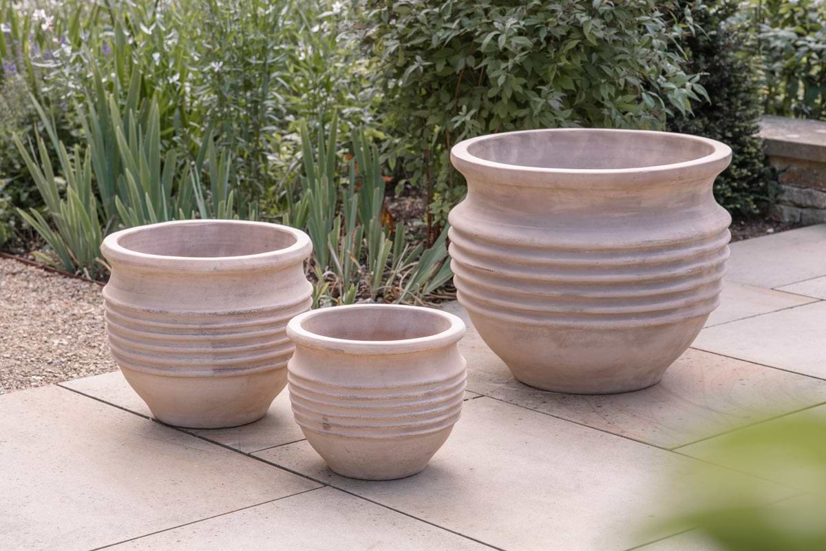 Three Ullu Terracotta Planters by nkuku, in natural terracotta with a ribbed design, sit on a stone patio amid lush greenery. These unglazed planters suit both indoor and outdoor use, adding rustic charm to any space.