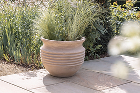 The nkuku Ullu Terracotta Planter sits outdoors on stone tiles, filled with tall ornamental grasses. Sunlight highlights the ribbed terracotta design as green plants and flowers provide a lush garden backdrop.