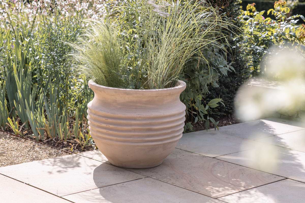 The nkuku Ullu Terracotta Planter sits outdoors on stone tiles, filled with tall ornamental grasses. Sunlight highlights the ribbed terracotta design as green plants and flowers provide a lush garden backdrop.