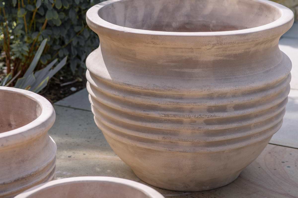 The nkuku Ullu Terracotta Planter – Terracotta, a large unglazed piece with a wide rim and five ridges, sits outdoors on stone. Nearby are similar planters for indoor and outdoor use, surrounded by green foliage.