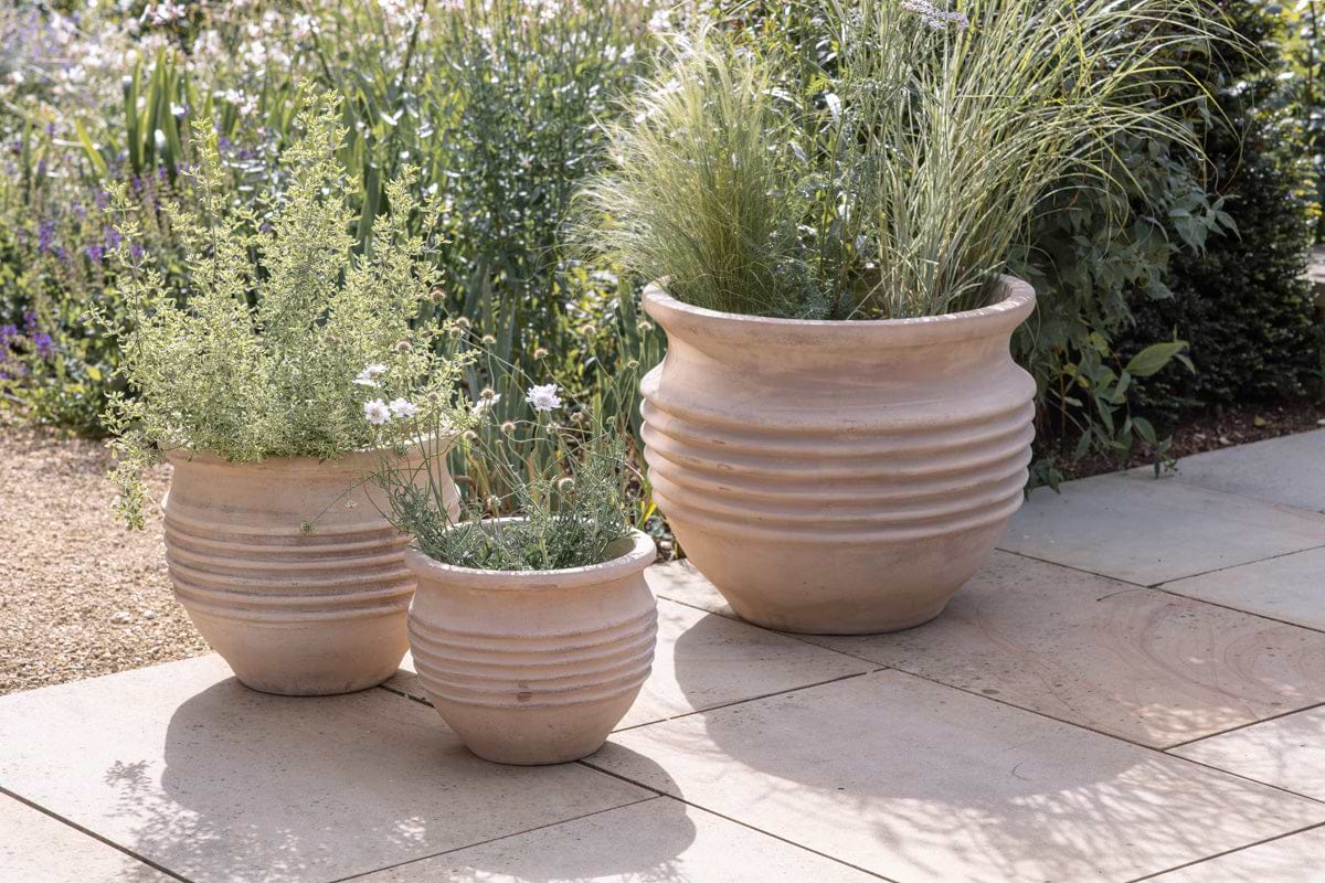 Three beige Ullu Terracotta Planters by nkuku, each with leafy green plants and white flowers, are arranged on light stone tiles in a sunlit garden with blurred greenery and flowers in the background.