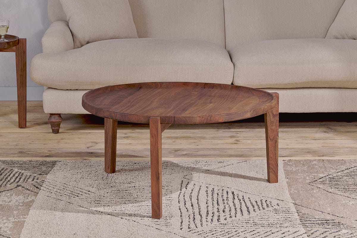 The Umar Mango Wood Coffee Table - Dark Brown by nkuku, featuring a circular design and three angular legs, is placed on a beige patterned rug in front of a light beige upholstered sofa. A wooden side table with a glass and book sits to the left.