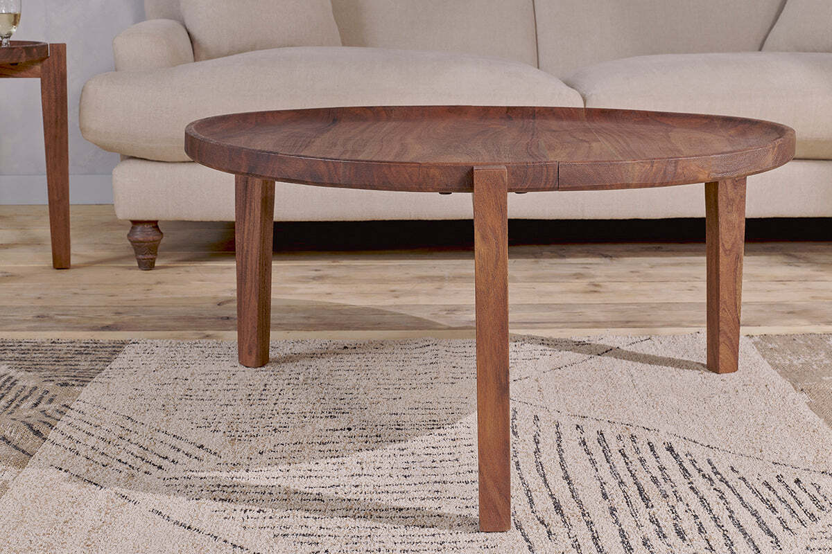 The Umar Mango Wood Coffee Table - Dark Brown by nkuku, featuring a sculptural circular design with three legs, is placed on a patterned beige rug in front of a light sofa, creating a cozy, modern living room setting.