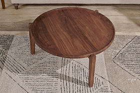 The nkuku Umar Mango Wood Coffee Table in dark brown, featuring a circular shape and tapered legs, sits on a rug with black and beige geometric patterns.