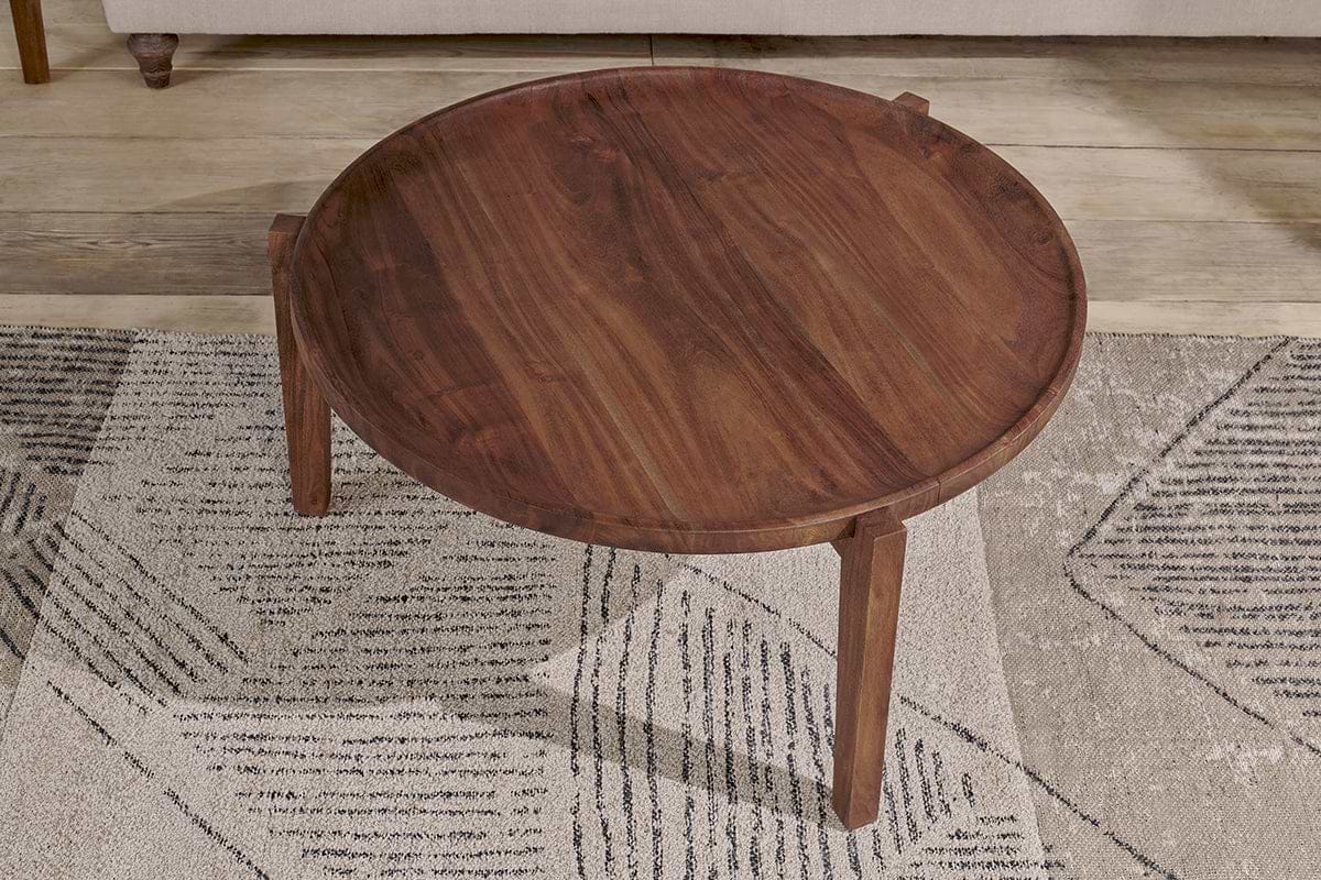 The nkuku Umar Mango Wood Coffee Table in dark brown, featuring a circular shape and tapered legs, sits on a rug with black and beige geometric patterns.