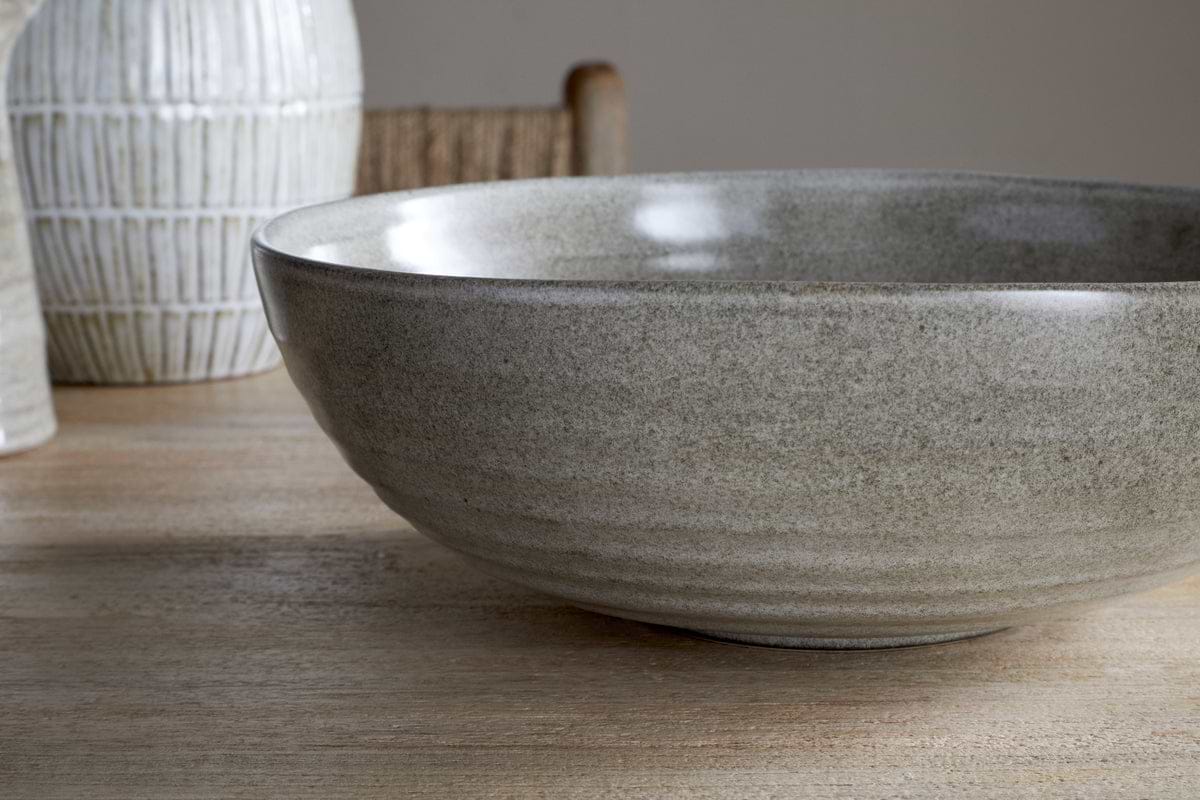 A close-up of the nkuku Valinha Earthenware Serving Bowl in grey, featuring a textured spiral design, sits on a wooden table with a white vase and woven chair softly visible in the background.