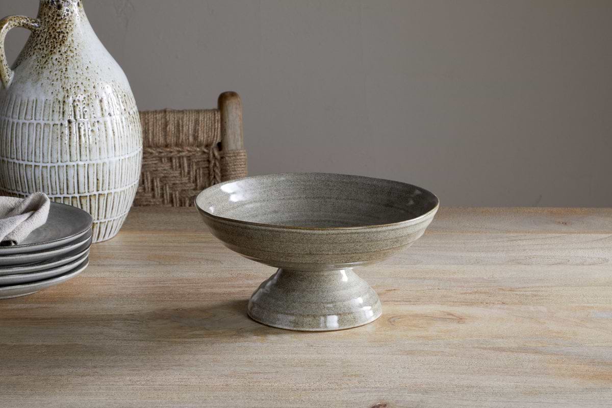 The nkuku Valinha Earthenware Footed Bowl in grey, with its speckled finish, rests on a wooden table beside plates, a napkin, and a jug—ideal for a rustic tablescape. A woven chair is visible in the background.