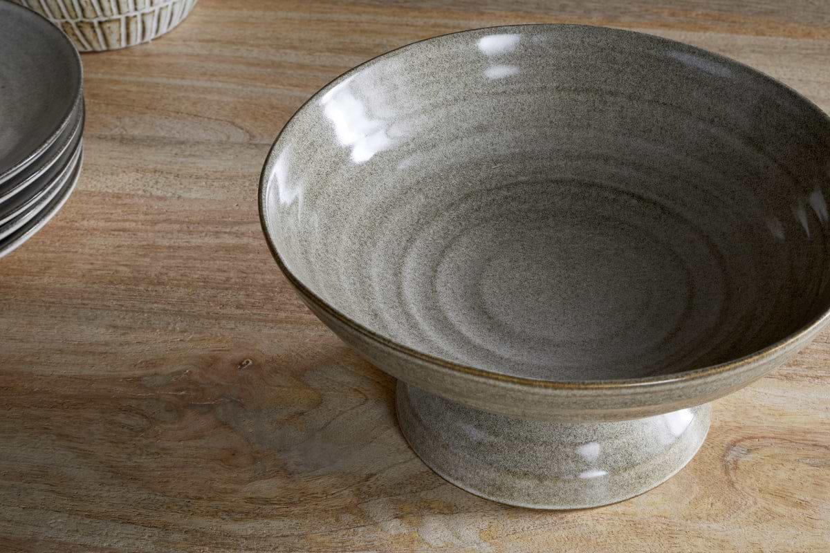 The nkuku Valinha Earthenware Footed Bowl - Grey, featuring a wide, shallow design and ribbed texture, adds rustic charm to any table setting. Shown on wood beside matching plates.
