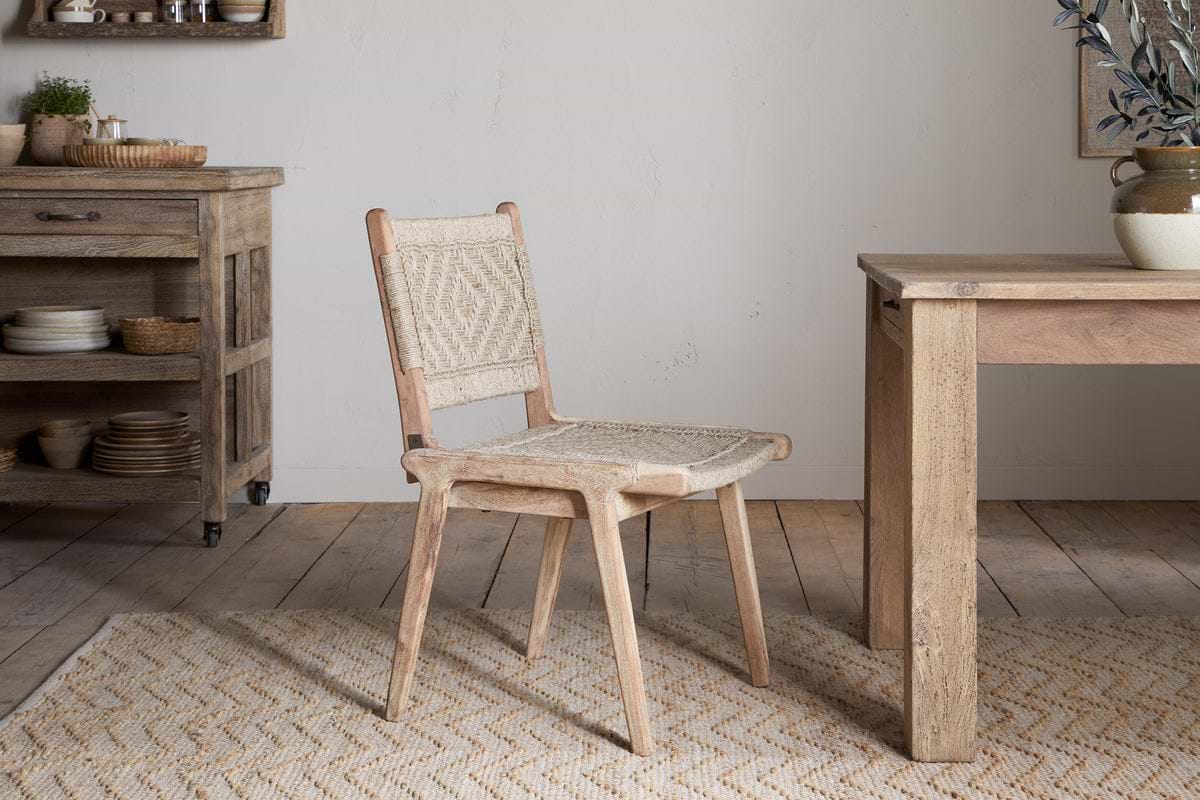 The nkuku Vinay Woven Armless Dining Chair in natural jute sits on a beige patterned rug in a bright, rustic dining room. Nearby are wooden shelves with dishes and baskets, and an acacia wood table with a vase of branches against a white wall.