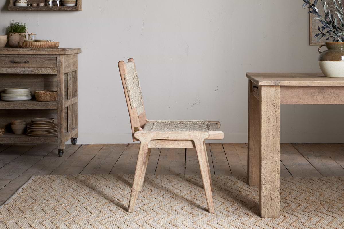 The Vinay Woven Armless Dining Chair by nkuku, made of acacia wood with a woven jute seat and backrest, is placed next to a wooden dining table in a rustic kitchen with wooden cabinets, shelves, and neutral decor.
