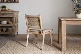 The nkuku Vinay Woven Armless Dining Chair in natural jute sits on a patterned rug in a rustic dining room with acacia wood furniture and neutral decor.