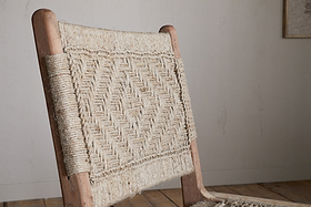 The Vinay Woven Armless Dining Chair by nkuku features acacia wood and a geometric diamond-patterned jute backrest. Set against a light wall and wooden floor, its natural style stands out in any dining space.