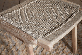 A close-up of the Vinay Woven Armless Dining Chair by nkuku, crafted from acacia wood with a geometric diamond-patterned jute seat, placed on a textured rug with a matching design.