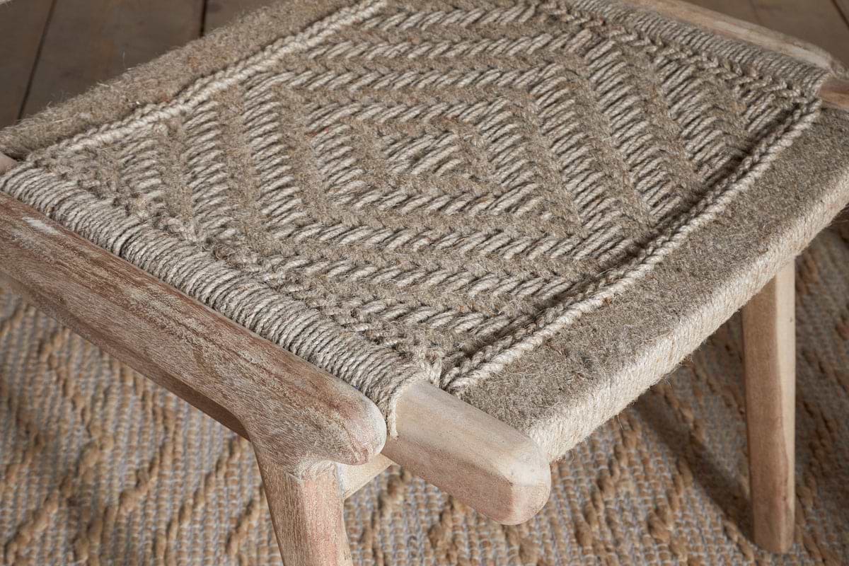 A close-up of the Vinay Woven Armless Dining Chair by nkuku, crafted from acacia wood with a geometric diamond-patterned jute seat, placed on a textured rug with a matching design.