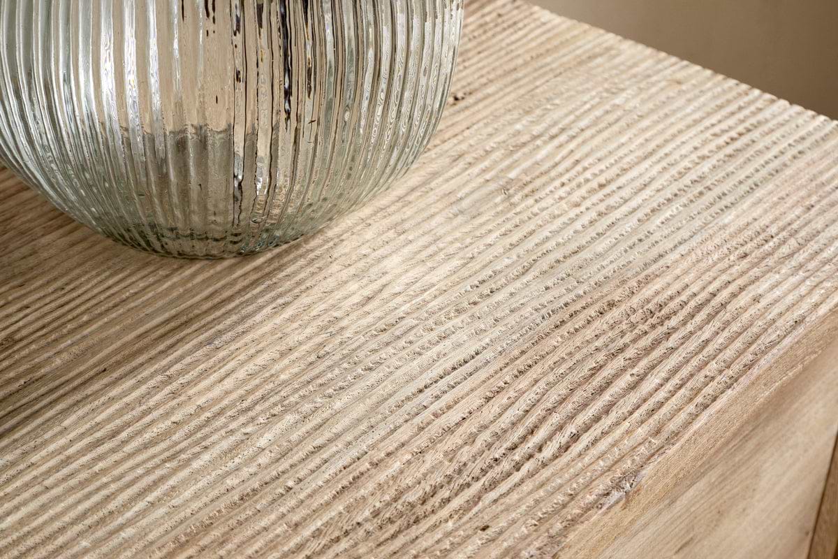 A close-up of a ribbed glass vase on a textured wooden surface, evoking the natural carved grooves of the nkuku Vivan Grooved Chest of Drawers.