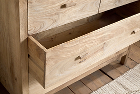 A close-up of the nkuku Vivan Grooved Chest of Drawers in natural finish, featuring one drawer partially open to reveal its wood grain. This stylish bedroom piece stands on a wooden floor beside a textured beige rug.