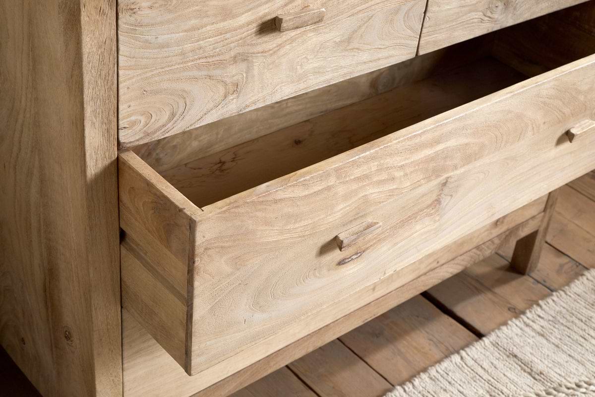 A close-up of the nkuku Vivan Grooved Chest of Drawers in natural finish, featuring one drawer partially open to reveal its wood grain. This stylish bedroom piece stands on a wooden floor beside a textured beige rug.