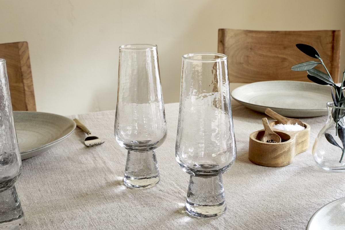 A close-up of a dining table set with two Visla Champagne Flutes by nkuku, grey ceramic plates, wooden chairs, a small wooden salt bowl with spoon, and a vase with green leaves on a beige tablecloth.